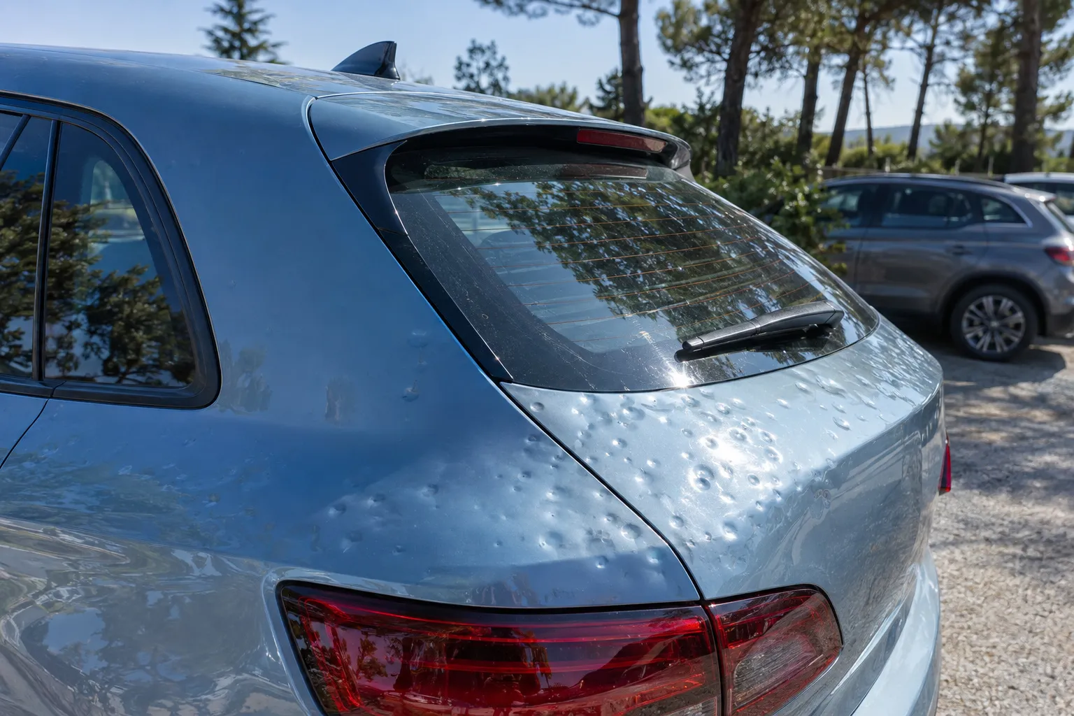 Coffre et lunette arrière d'une compacte bleu clair marqués de petits impacts de grêle, sur un parking en plein air.